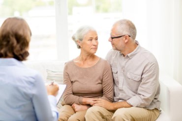 an older couple sit holding hands on a couch while a woman makes notes on a clipboard in the foreground