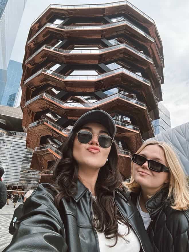 two young women in sunglasses pose for a selfie in front of an abstract building