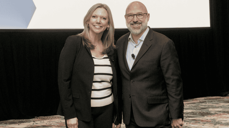 an image of a smiling blond woman in a black blazer and black and white striped shirt standing next to a smiling bearded bald man in glasses and a dark grey blazer and checkered shirt