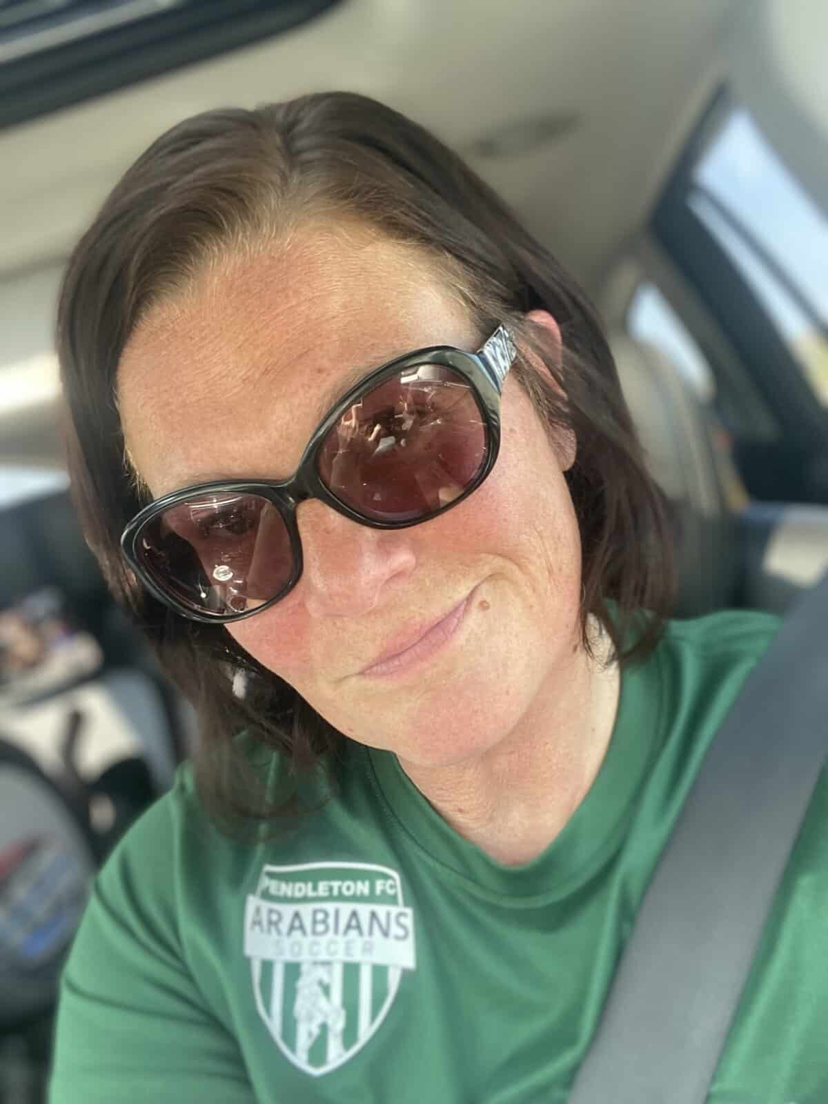 a woman in a green shirt with short dark hair and sunglasses smiles at the camera