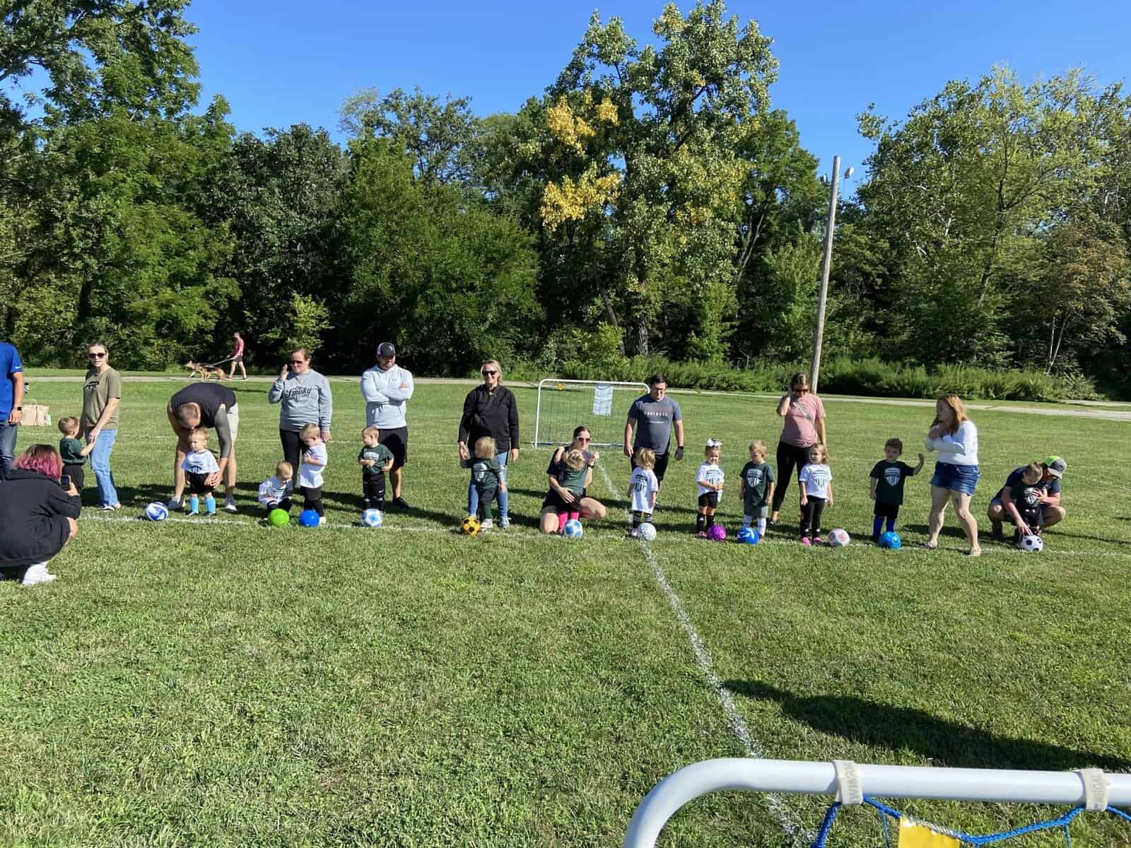 children and their parents stand in a line in front of soccer balls, presumably at a soccer camp